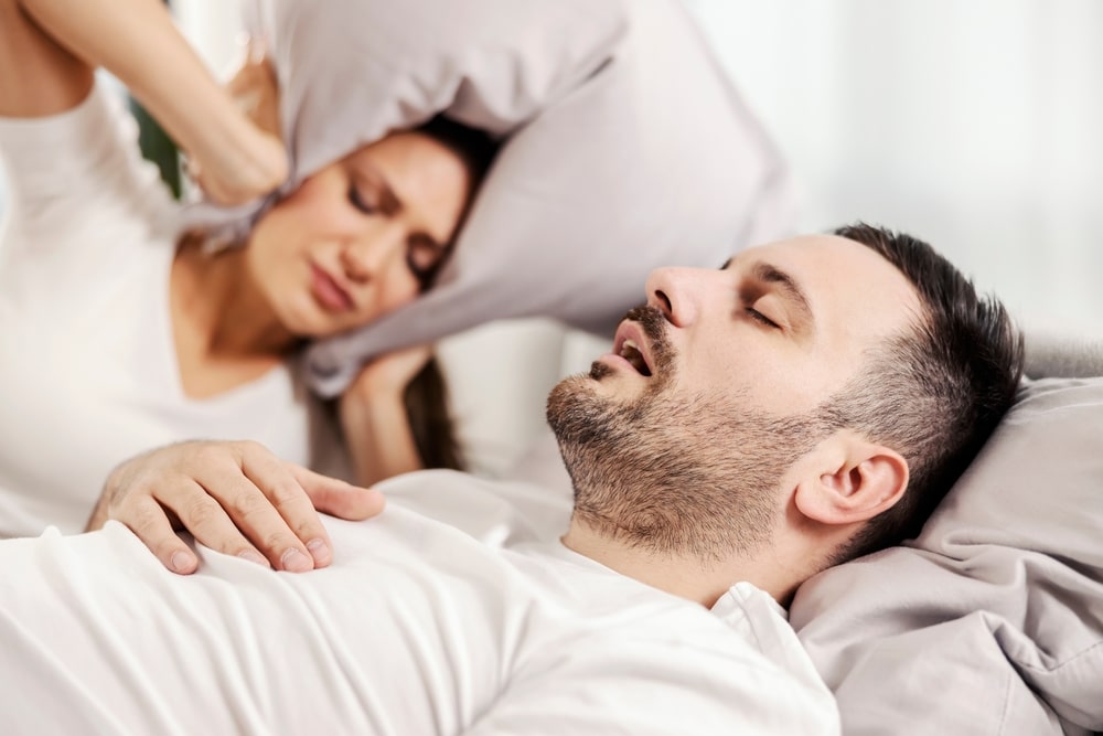 Man snoring in bed next to his wife who cannot fall asleep due to the noise.